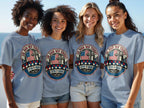 Four women smiling in navy t-shirts with "In God We Trust" and 1776+2026 graphic.