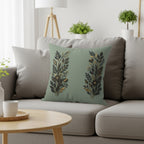 Sage green throw pillow with gold and black floral design on a gray sofa.