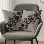 Two beige throw pillows with black floral vine designs on a grey armchair.
