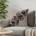 Beige throw pillow with black and gold botanical design on a grey couch.