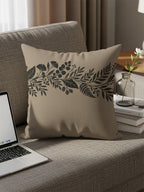 Beige throw pillow with black botanical design on a sofa with a laptop.