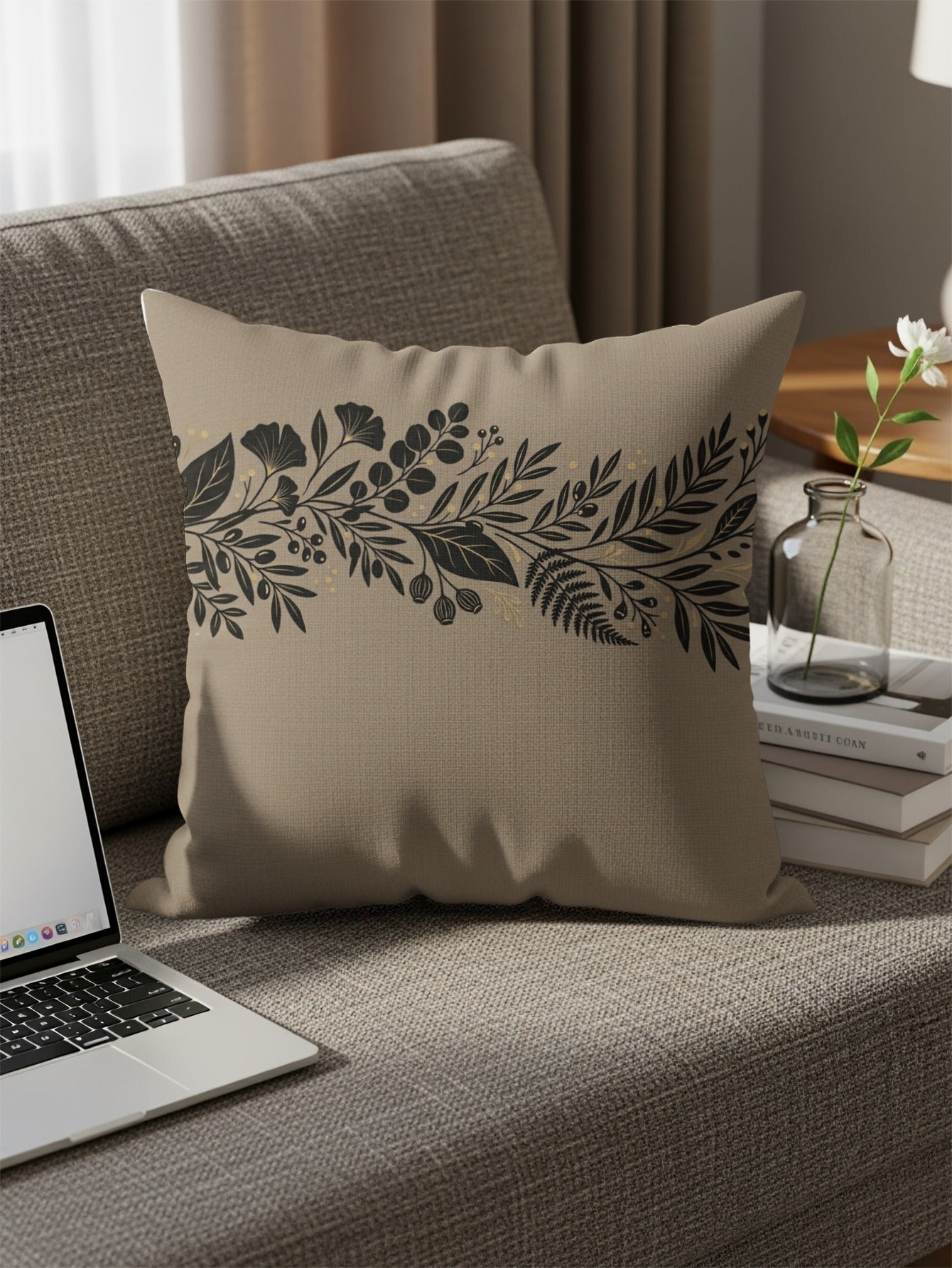 Beige throw pillow with black botanical design on a sofa with a laptop.