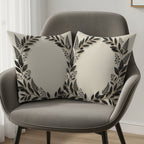 Two cream throw pillows with black botanical wreath designs on a grey chair.