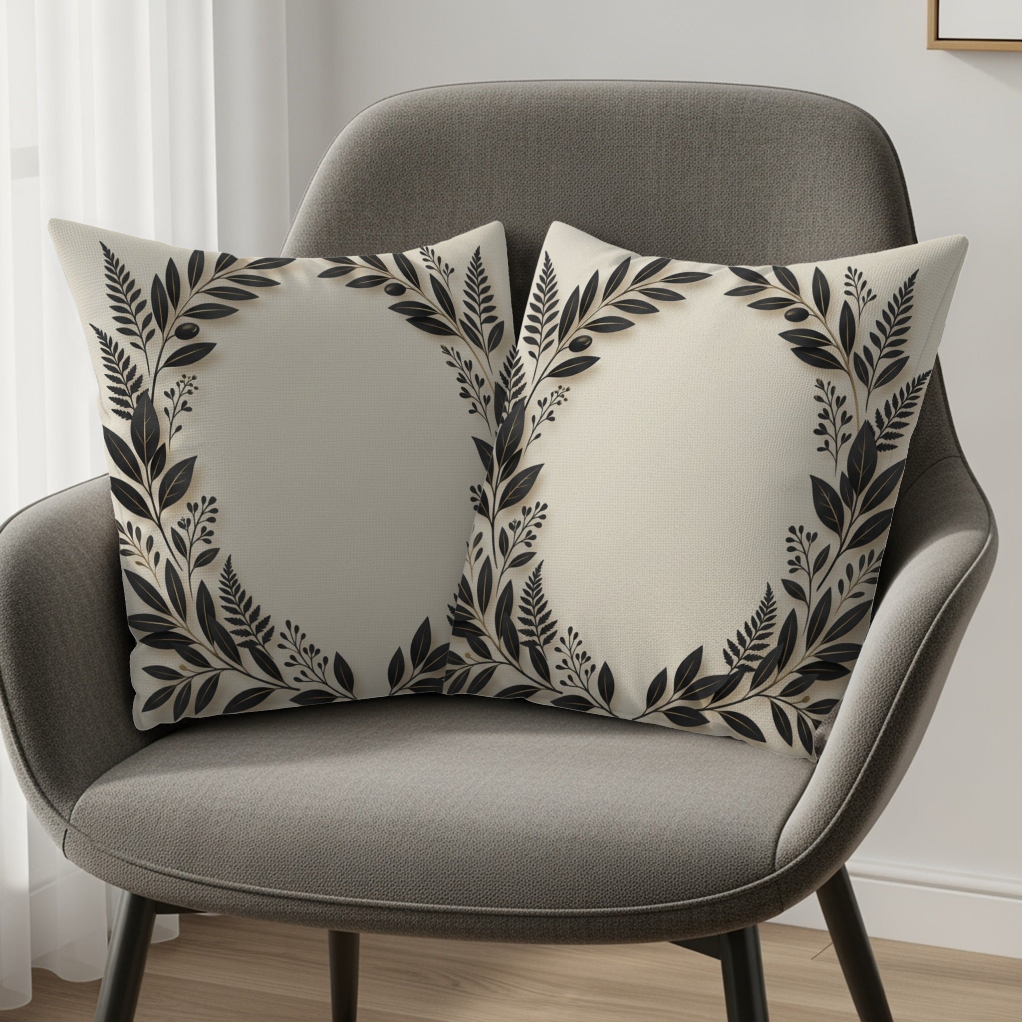 Two cream throw pillows with black botanical wreath designs on a grey chair.