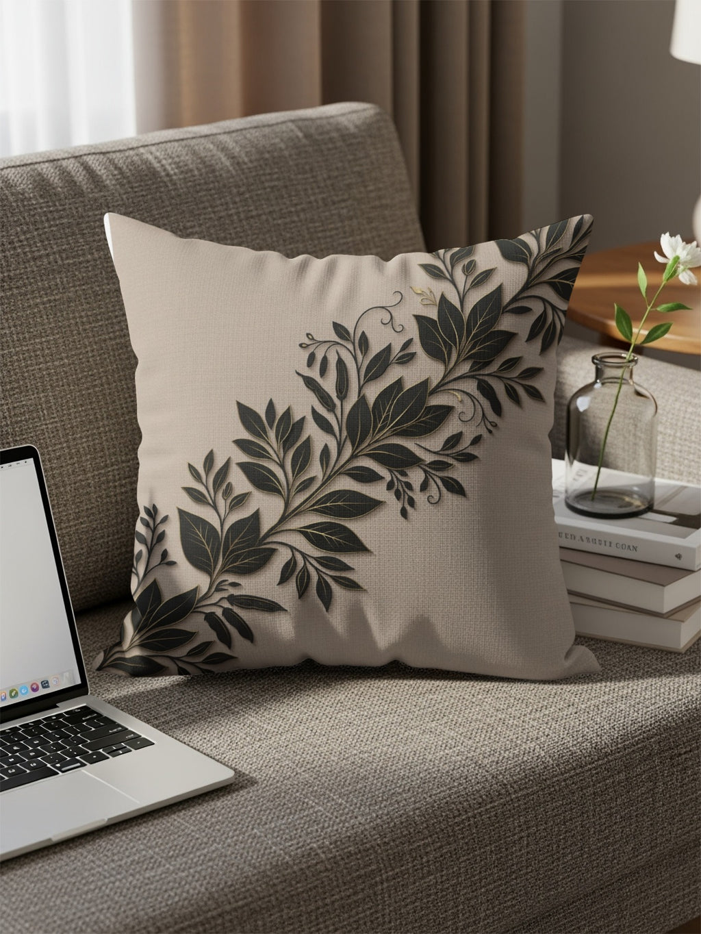 Beige throw pillow with dark floral vine pattern on sofa with laptop and vase.