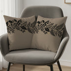 Two taupe throw pillows with black and gold floral border design on gray chair.