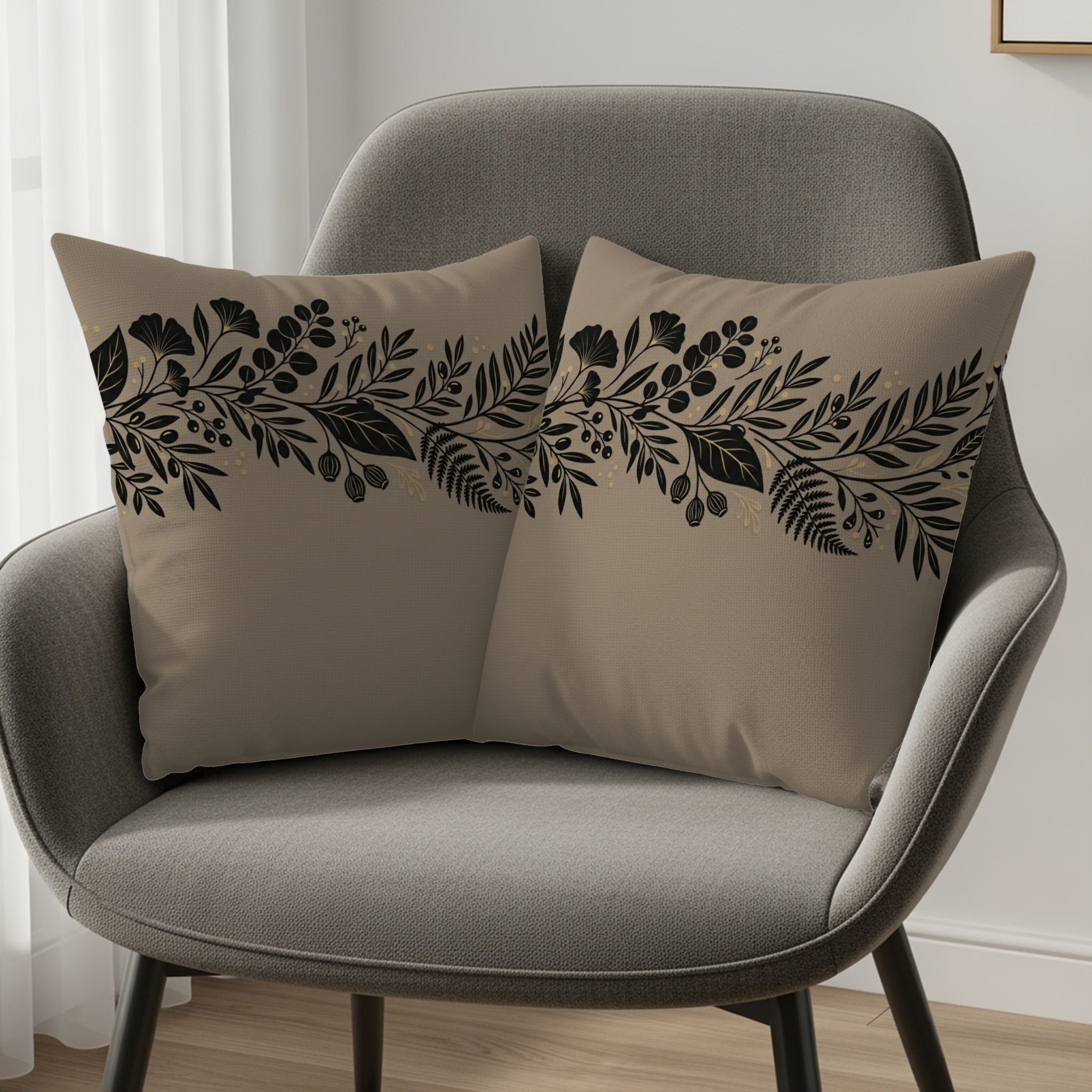 Two taupe throw pillows with black and gold floral border design on gray chair.
