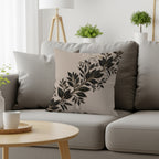 Beige throw pillow with an ornate black leaf vine design on a gray sofa.