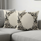 Two cream decorative pillows with black floral wreath pattern on a sofa.